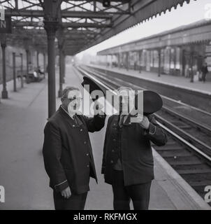 1968, deux employés de la station ferroviaire britannique en uniforme, un contremaître et contrôleur debout ensemble sur la plate-forme à la gare de Blackheath, soulever leur casquette, Blackheath, Londres, Angleterre, Royaume-Uni. Banque D'Images