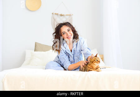 Portrait of attractive smiling woman in pyjama avec Bengal chat posant sur le lit chez lui Banque D'Images