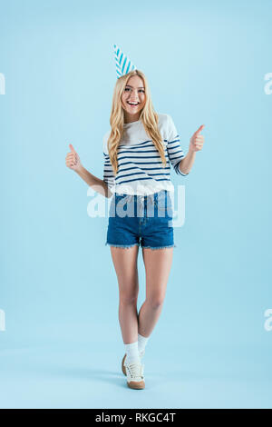 Studio shot of attractive blonde girl in party hat posing with Thumbs up sur fond bleu Banque D'Images