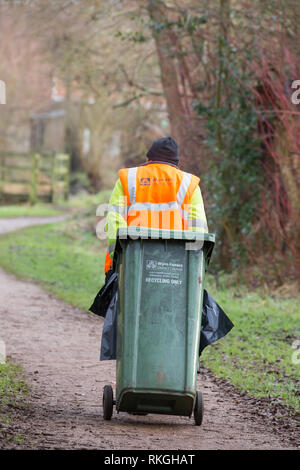 Vue arrière d'un employé du conseil masculin dans une veste haute visibilité et un chapeau tirant la poubelle verte de recyclage derrière lui dans un parc public. UK bin Man au travail. Banque D'Images