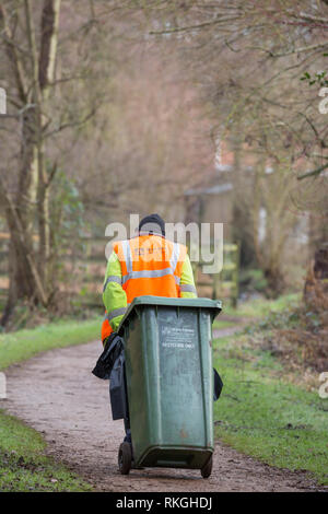 Vue arrière d'un employé du conseil dans une veste haute visibilité tirant la poubelle de recyclage verte derrière lui dans un parc public. UK bin man, bin men au travail. Banque D'Images