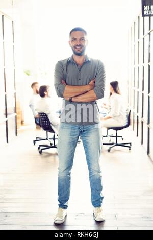 Full Length portrait of smiling young man standing in office. Creative male executive au démarrage avec des gens qui travaillent en arrière-plan Banque D'Images