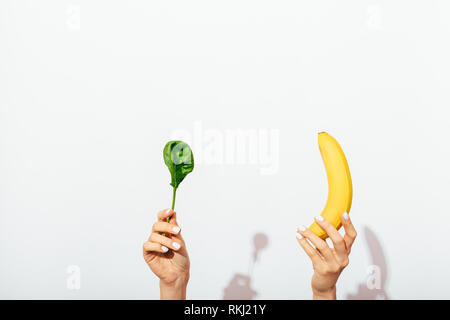 Les mains de femmes avec des épinards en feuilles et manucure tenant une banane sur fond blanc, d'ombre. Banque D'Images