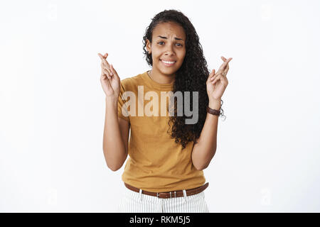 Studio shot of silly nerveux et jeune femme à la peau sombre avec des cheveux bouclés crossing soulevées doigts souhaiter bonne chance, fronçant et souriant inquiet Banque D'Images