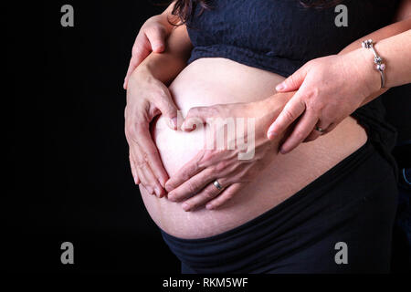 Femme enceinte avec des mains du mari sur son ventre en forme de coeur symbolisant l'amour. Isolé sur fond noir. Banque D'Images