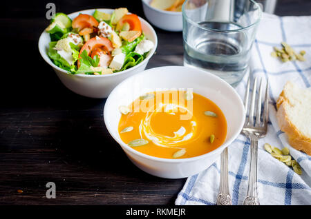 Servant pour le déjeuner, de soupe au potiron dans bol blanc et vert clair frais salade de légumes et d'eau en verre sombre sur fond de bois rustique, na Banque D'Images