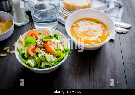 Servant pour le déjeuner, de soupe au potiron dans bol blanc et vert clair frais salade de légumes et d'eau en verre sombre sur fond de bois rustique, na Banque D'Images