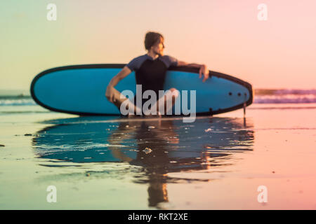 Silhouette de surf homme assis avec un surf sur le bord de la plage à l'heure du coucher du soleil Banque D'Images