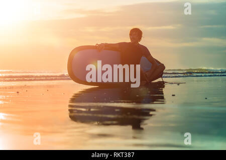 Silhouette de surf homme assis avec un surf sur le bord de la plage à l'heure du coucher du soleil Banque D'Images