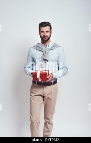 Présent pour vous. Homme élégant dans des vêtements décontractés tient une boîte cadeau et smiling at camera while standing en studio sur un fond gris. Close-up portrait Banque D'Images