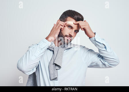 Oops...jeune homme barbu en vêtements de détente est de mettre des cataplasmes sur son front et à la caméra à tout en se tenant sur un fond gris. Concept de la douleur Banque D'Images