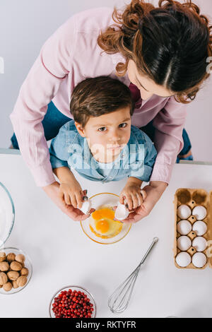 Vue de dessus de maman avec peu de sérieux fils casser des œufs dans le bol sur fond clair Banque D'Images