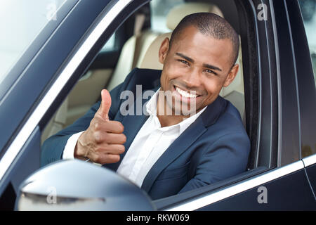 Young african american businessman chauffeur assis à l'intérieur de la voiture à la recherche ou la fenêtre showing thumb up smiling smiling close-up Banque D'Images