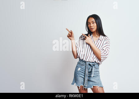 Regardez là-bas. Cute asian woman with long hair pointant vers le haut et souriant tout en se tenant sur un fond gris. Close up portrait. Banque D'Images