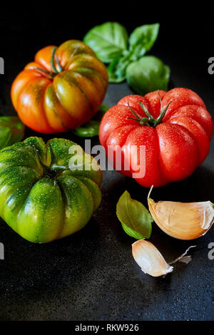 Rouge, vert et orange les tomates scintillants de gouttelettes d'eau sur une surface réfléchissante sombre avec des gousses d'ail et basilic, avant-plan copy space Banque D'Images