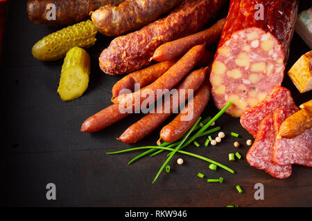 Assortiment de saucisses épicées chevronnés spécialisés organisés dans le coin sur un fond sombre avec la ciboulette, poivre, des concombres ou cornichons un Banque D'Images