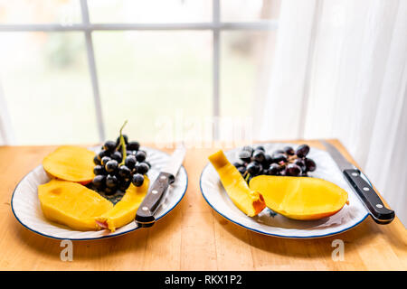 Libre de couper les tranches de mangue sur les moitiés de fruits deux plaques sur table en bois par fenêtre avec des raisins noirs et des couteaux pour petit-déjeuner romantique ou un en-cas Banque D'Images