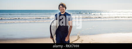 Beau jeune sportif posant avec sa planche de surf sous le bras dans sa combinaison sur une plage tropicale de sable long format bannière Banque D'Images