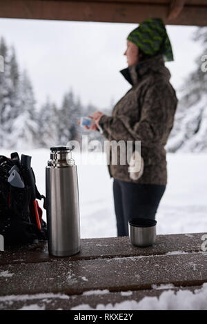 Jeune femme en matériel de randonnée debout dans un refuge de boire du café tout en faisant une pause à partir d'une randonnée dans une forêt d'hiver Banque D'Images