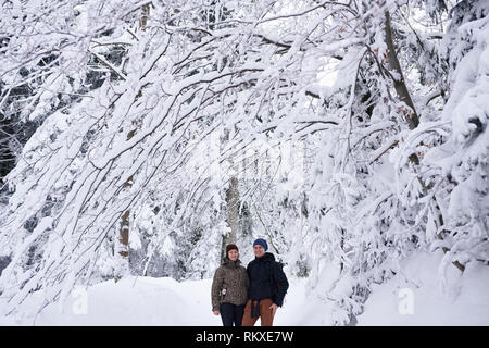 Portrait of a smiling young couple in hiking gear standing arm in arm ensemble sur un sentier de la forêt enneigée en hiver Banque D'Images