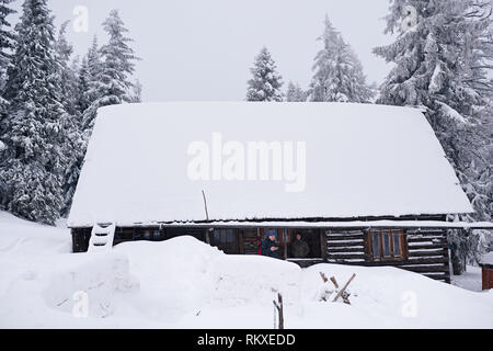 Jeune couple debout sur le porche d'un chalet en bois couvert de neige dans une forêt en plein hiver Banque D'Images