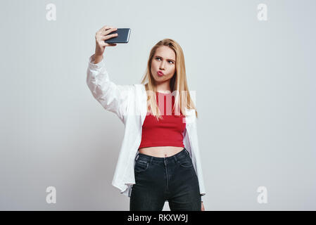 Temps selfies. Jolie jeune femme avec des cheveux longs en selfies son smartphone et faire une grimace en se tenant debout à l'arrière-plan gris Banque D'Images