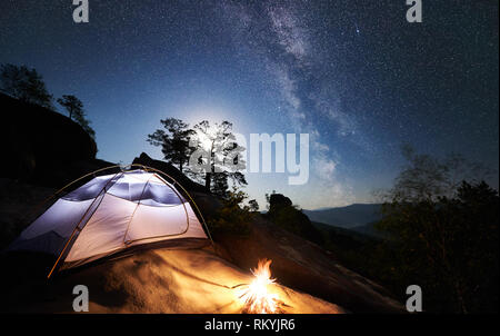 Camping touristique à une nuit d'été sur rocky mountain. Camp sous tente et rayonnante nuit incroyable ciel plein d'étoiles et de Voie lactée. Sur le fond beau ciel étoilé, gros rochers et arbres Banque D'Images