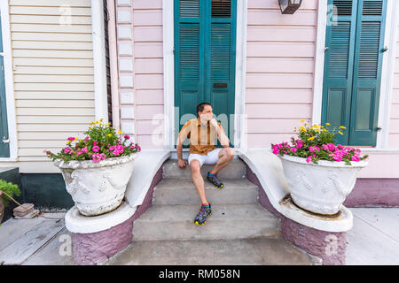 Jeune homme assis smiling happy sur mesures escaliers porche dans la Nouvelle-Orléans par colorful pink blue green architecture porte avec des fleurs jaunes en Banque D'Images