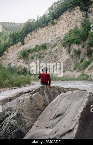 Jeune homme en veste rouge et noir hat se dresse sur un rocher et bénéficie d'mountain nature. Montagnes épique sur l'arrière-plan. Concept de vie en plein air dans la nature Banque D'Images