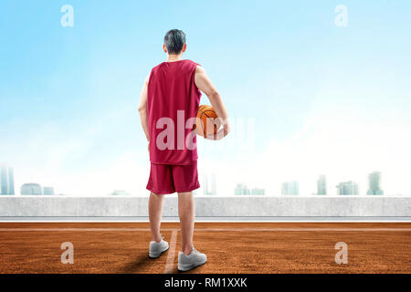 Vue arrière de l'asian man in red sportswear tenant le basket-ball debout sur la piscine en plein air à la cour de basket-ball à la ville avec fond de ciel bleu Banque D'Images