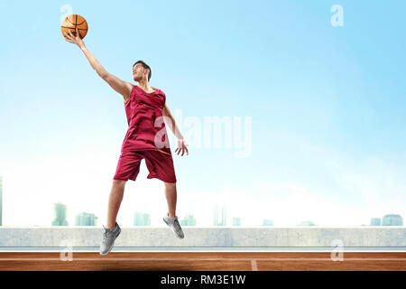 Bel asiatique homme basket-ball player jumping high et rebondir la balle pour marquer dans l'air extérieur de basket-ball avec des paysages urbains et de fond de ciel bleu Banque D'Images