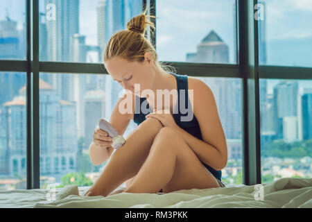 Jeune femme nitae sur le lit chez lui et faire l'épilation avec l'épilateur sur les jambes. Sur l'arrière-plan d'une fenêtre donnant sur la grande ville, des gratte-ciel Banque D'Images