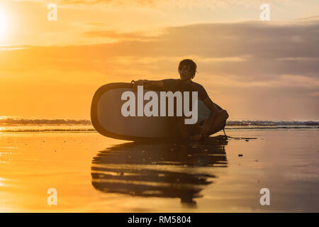 Silhouette de surf homme assis avec un surf sur le bord de la plage à l'heure du coucher du soleil Banque D'Images