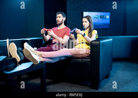 Jeune couple de jeux vidéo avec consoles de jeu assis sur le canapé dans la pièce sombre de la club de lecture Banque D'Images