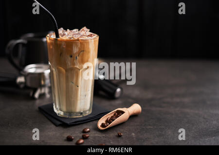 Café crème avec de la glace dans un grand verre et les grains de café, portafilter, infalsifiable et pot à lait sur le béton sombre sur fond de table en bois noir. Somme à froid Banque D'Images