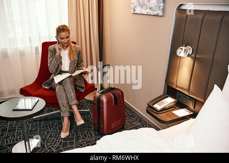 Femme appelant en voyage d'affaires. Jeune et élégant smartphone et businesswoman with suitcase sitting on sofa à chambre d'hôtel. Tablette numérique à la table Banque D'Images