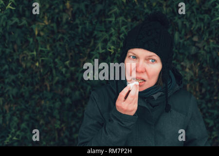 Femme d'appliquer le baume de lèvre à la rue par une froide journée d'hiver contre le mur de lierre commun Banque D'Images