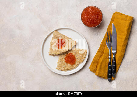 Crêpes de fête avec du caviar rouge sur plaque blanche avec une fourchette, un couteau et une serviette, pot de caviar, sur fond blanc. Concept de repas de fête. Vue depuis l'ab Banque D'Images