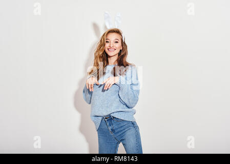 Portrait de jeune fille enjouée, pétillante portant des jeans, pull bleu et oreilles de lapin se tenant la main comme le lièvre, smiling at camera, isolé sur fond blanc. Banque D'Images