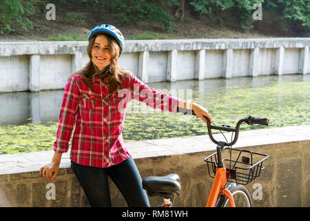 Mode de transport écologique en location. Belle jeune femme kasazy portant un casque bleu et cheveux longs pose debout à côté d'un orange-colore Banque D'Images