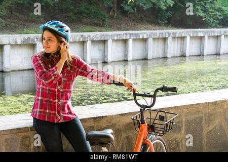 Mode de transport écologique en location. Belle jeune femme kasazy portant un casque bleu et cheveux longs pose debout à côté d'un orange-colore Banque D'Images