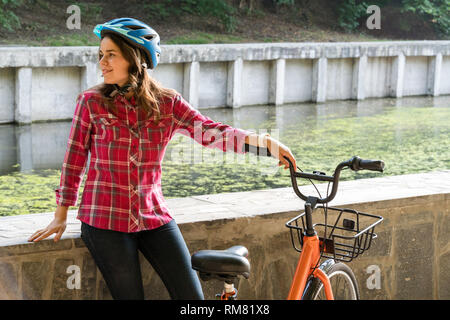 Mode de transport écologique en location. Belle jeune femme kasazy portant un casque bleu et cheveux longs pose debout à côté d'un orange-colore Banque D'Images