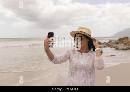 Jeune femme avec chapeau et des lunettes en tenant avec selfies mobile phone on beach Banque D'Images
