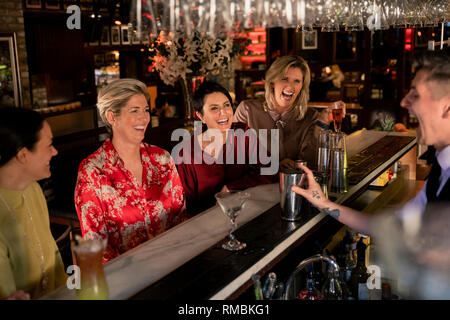 Petit groupe de femmes mûres bénéficiant d'une nuit, assis au bar. Ils rient avec le jeune barman qui fait un cocktail. Banque D'Images
