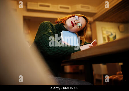 Rêver de quelque chose de beau. Belle jeune femme gaie redhaired en lunettes, pull en laine chaud vert, en utilisant son téléphone et ordinateur portable, tout en Banque D'Images