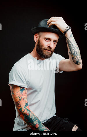 Hipster barbu joyeux en chapeau et t-shirt avec des tatouages sur les bras sur fond noir Banque D'Images