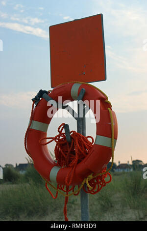 Bouée orange attachée à un câble sur un post sur la plage avec l'herbe et des maisons de plus en plus floue dans l'arrière-plan. Banque D'Images