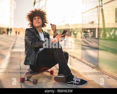 Beau jeune homme noir avec coiffure afro à écouter de la musique avec un téléphone intelligent dans une ville ensoleillée Banque D'Images