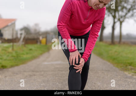 Femme jogger serrant son genou blessé avec une grimace de douleur alors qu'elle fait une pause au cours de son entraînement sur une route de campagne en hiver dans un close up portrait Banque D'Images