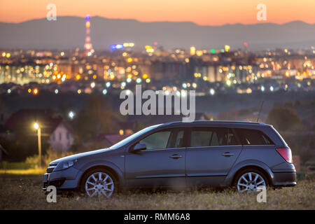 Gray voiture garée dans la nuit dans les prés verts sur fond de lumières de ville loin des immeubles et dark mountain ridge sous ciel clair au coucher du soleil. Transp Banque D'Images